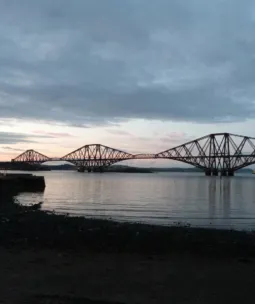 Forth Bridge at dusk – iconic red cantilever railway bridge in Scotland spanning the Firth of Forth, with steel triangular framework reflected in calm waters and a rocky shoreline in the foreground.