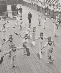 Rows of children on the deck of a ship, holding flags doing semaphore
