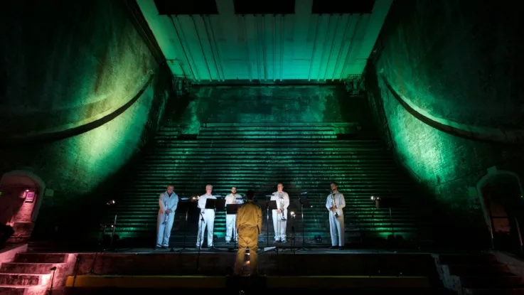 A concert in the bascule chambers at Tower Bridge. The walls are lit in green, as five musicians are led by a conductor.